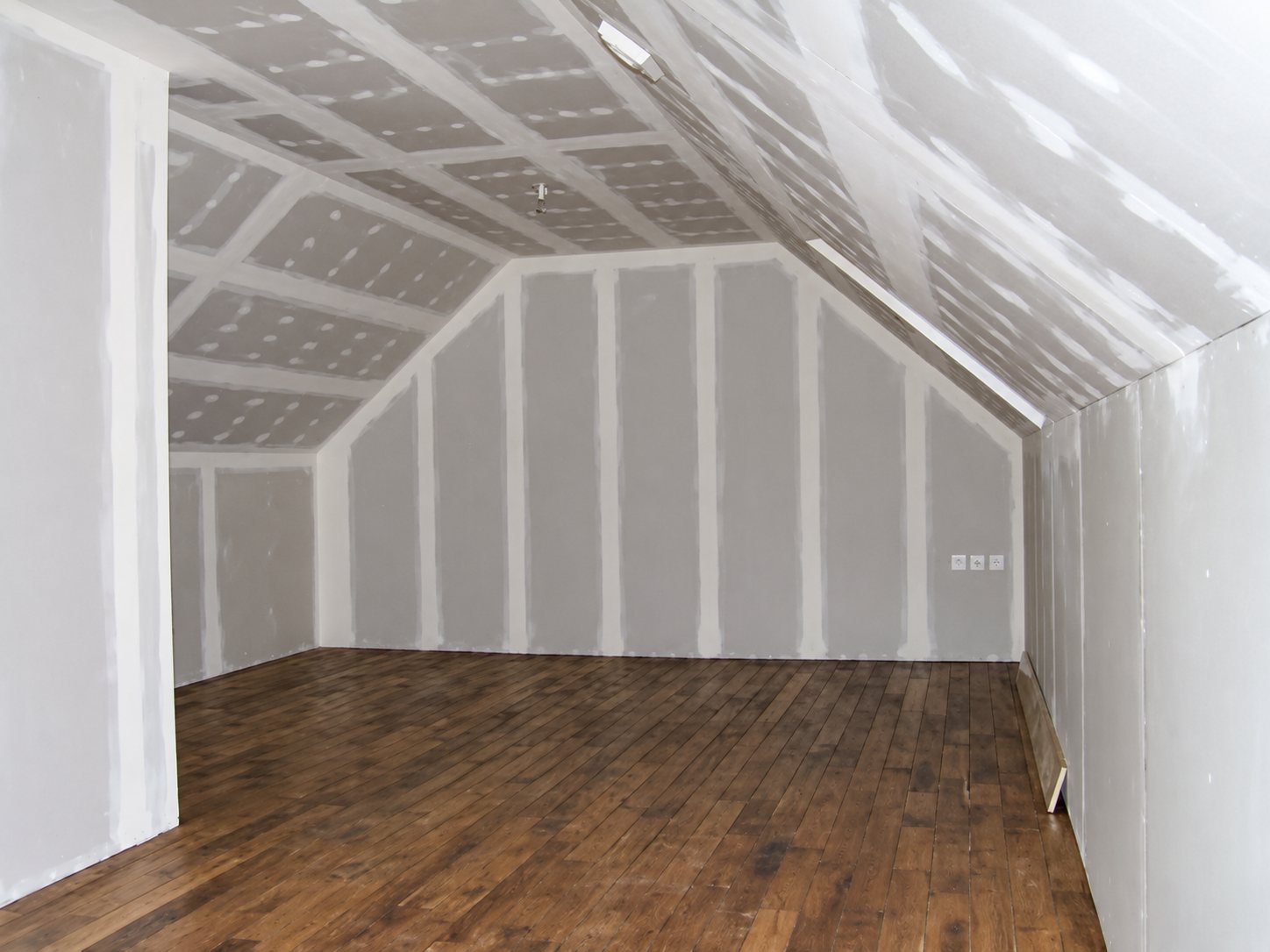 Un grand comble sous toiture dont les murs et le plafond sont recouverts de plaques de plâtre aux joints enduits, avec un parquet en bois foncé au sol.