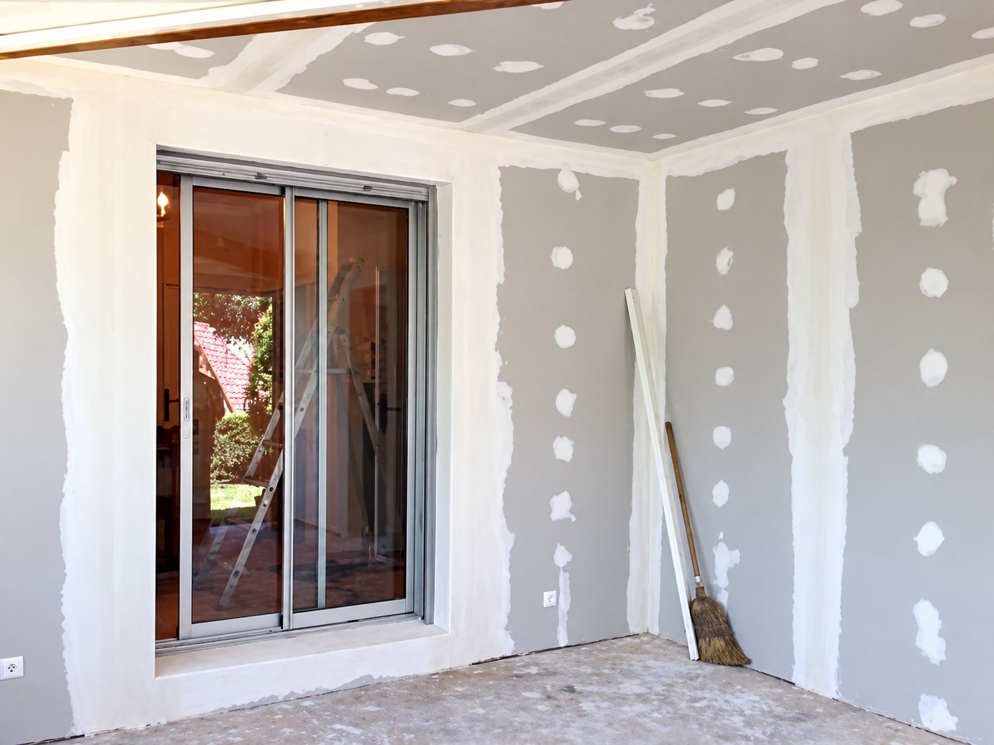 Pièce en cours de rénovation dont les murs et le plafond en plaques de plâtre sont jointoyés, avec une baie vitrée coulissante et un balai posé contre le mur.