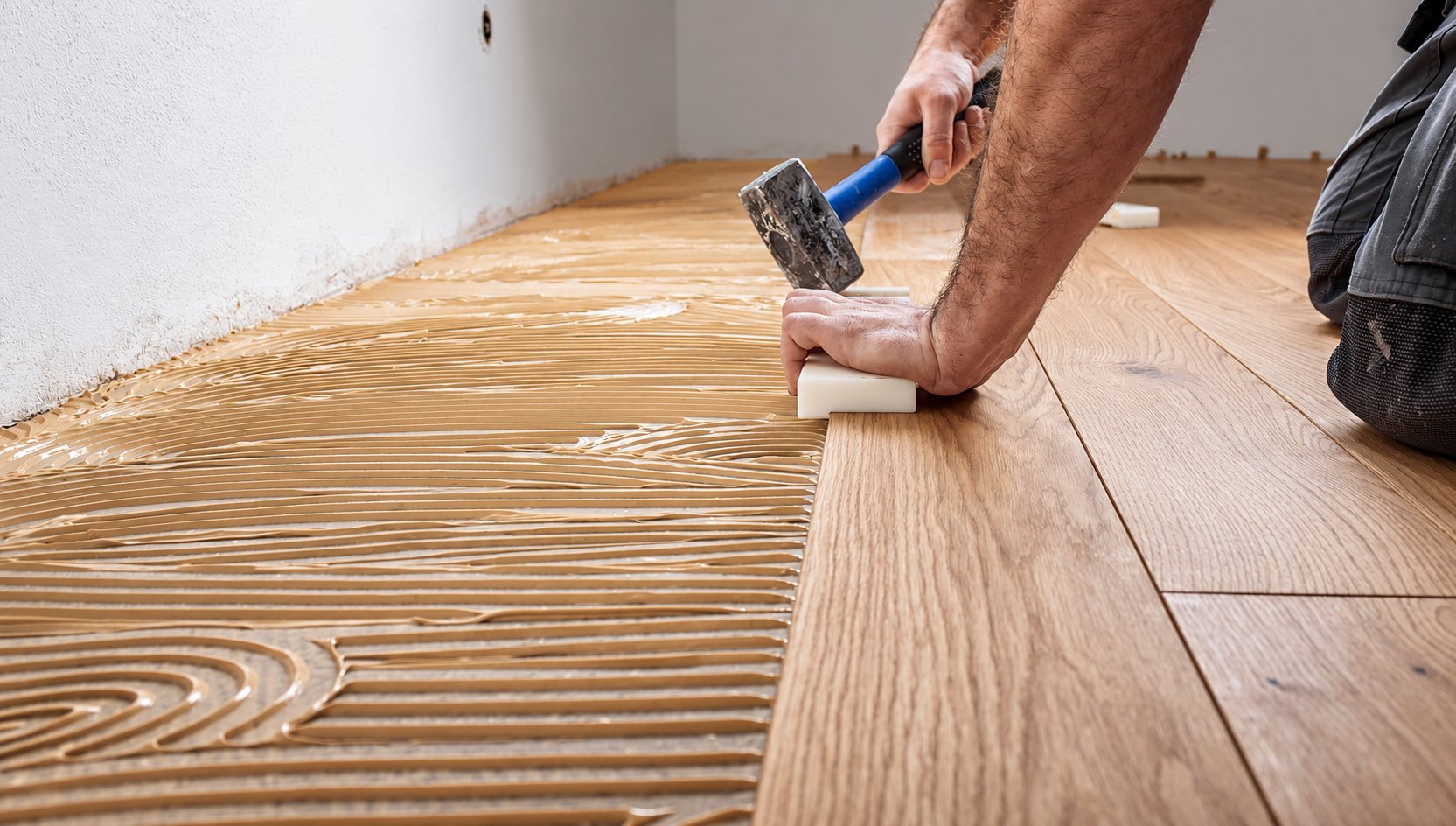 Un ouvrier à genoux tape avec un maillet sur une cale pour poser des lames de parquet en chêne sur une colle étalée au sol.
