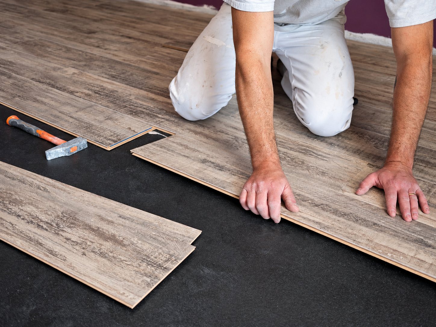 Un poseur en vêtements blancs, à genoux, assemble des lames de parquet stratifié gris sur un support sombre, avec un marteau posé à côté.