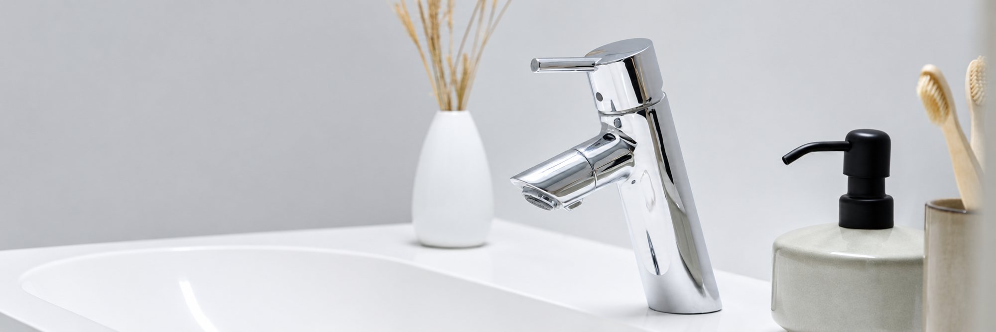 Un mélangeur chromé posé sur un lavabo blanc, accompagné d'un distributeur de savon noir, de brosses à dents en bambou et d'un vase avec des herbes séchées.