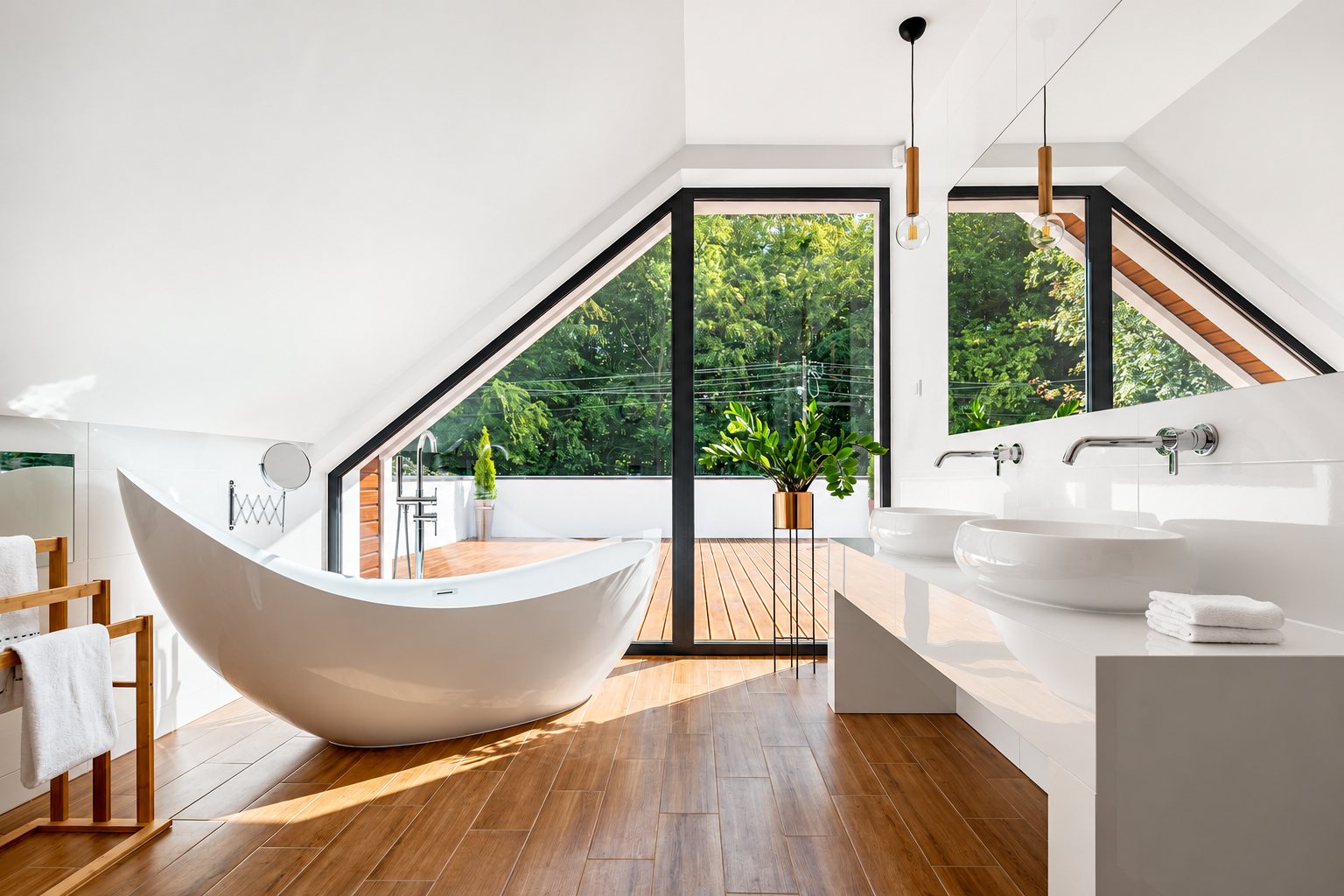 Salle de bain lumineuse sous les combles avec une baignoire îlot blanche, deux vasques sur un plan blanc, un parquet effet bois et une grande baie vitrée ouvrant sur une terrasse et des arbres.