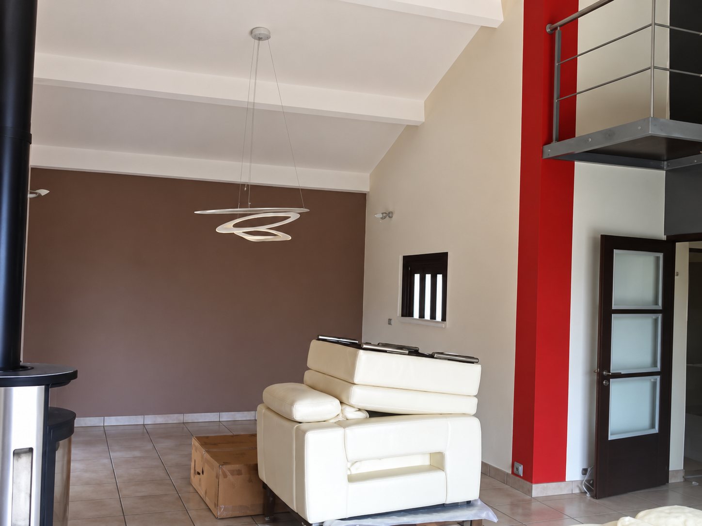 Un salon spacieux aux murs taupe et blancs avec une bande rouge verticale, un canapé en cuir blanc, un luminaire suspendu design et une mezzanine à garde-corps métallique.