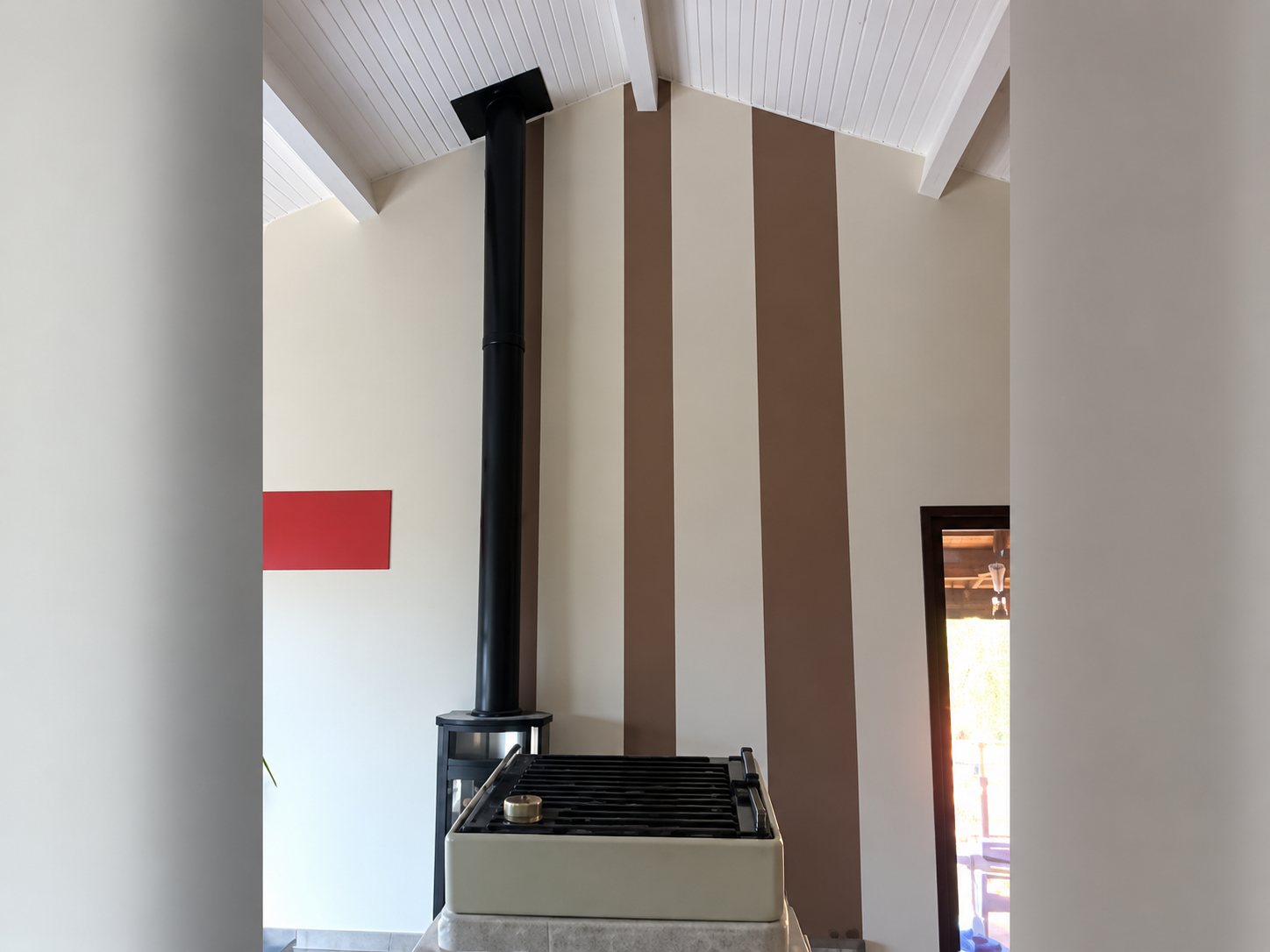 Intérieur d'une pièce sous plafond lambrissé blanc avec un poêle à bois noir et des bandes marron peintes sur le mur.